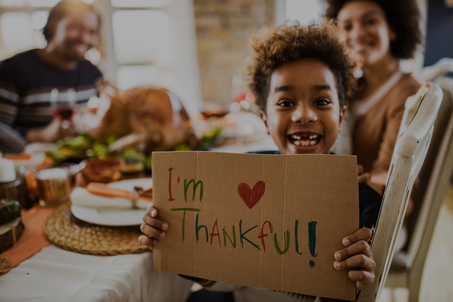A boy at a family holiday table holds up a handwritten sign that reads “I’m Thankful!”