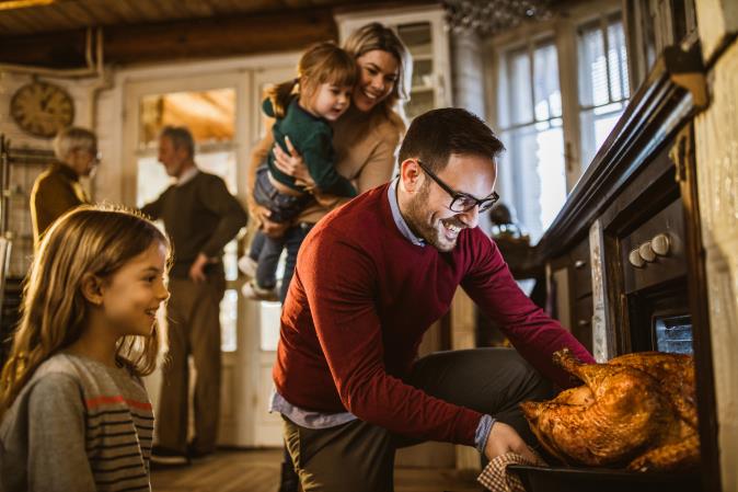 A man in a red sweater checks a turkey in the oven while his family watches. Open larger view.